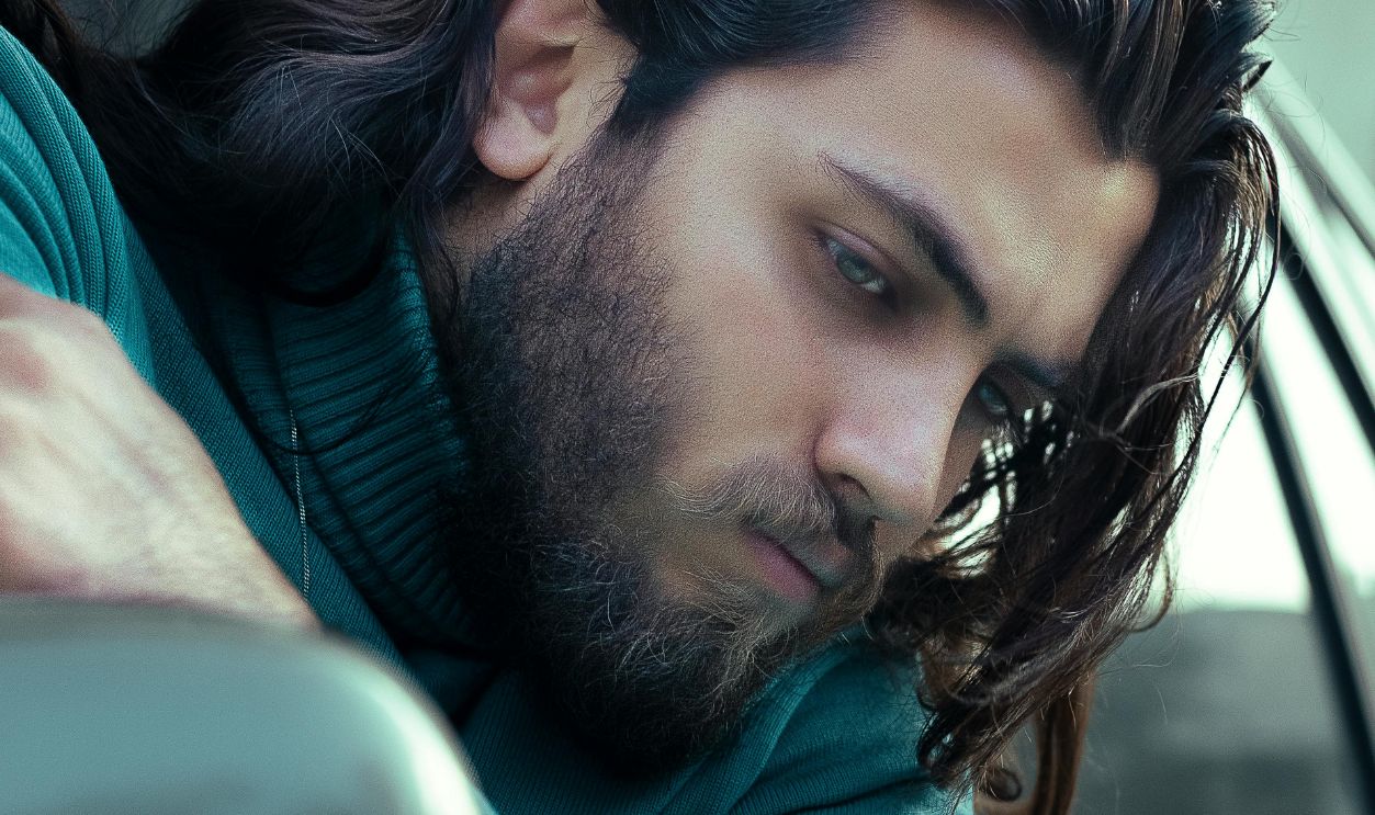 Thoughtful man in teal turtleneck looks out from car window with long hair flowing