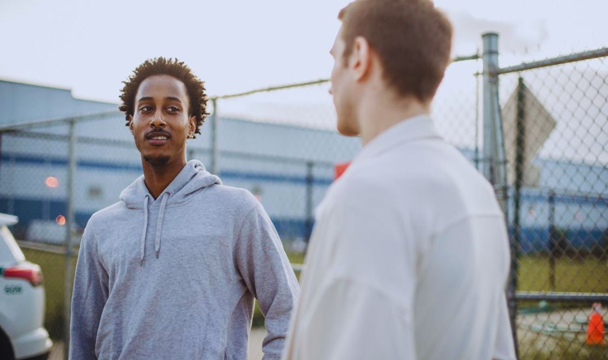 Two young men engaging in conversation near a fence outdoors with cars and buildings in the background
