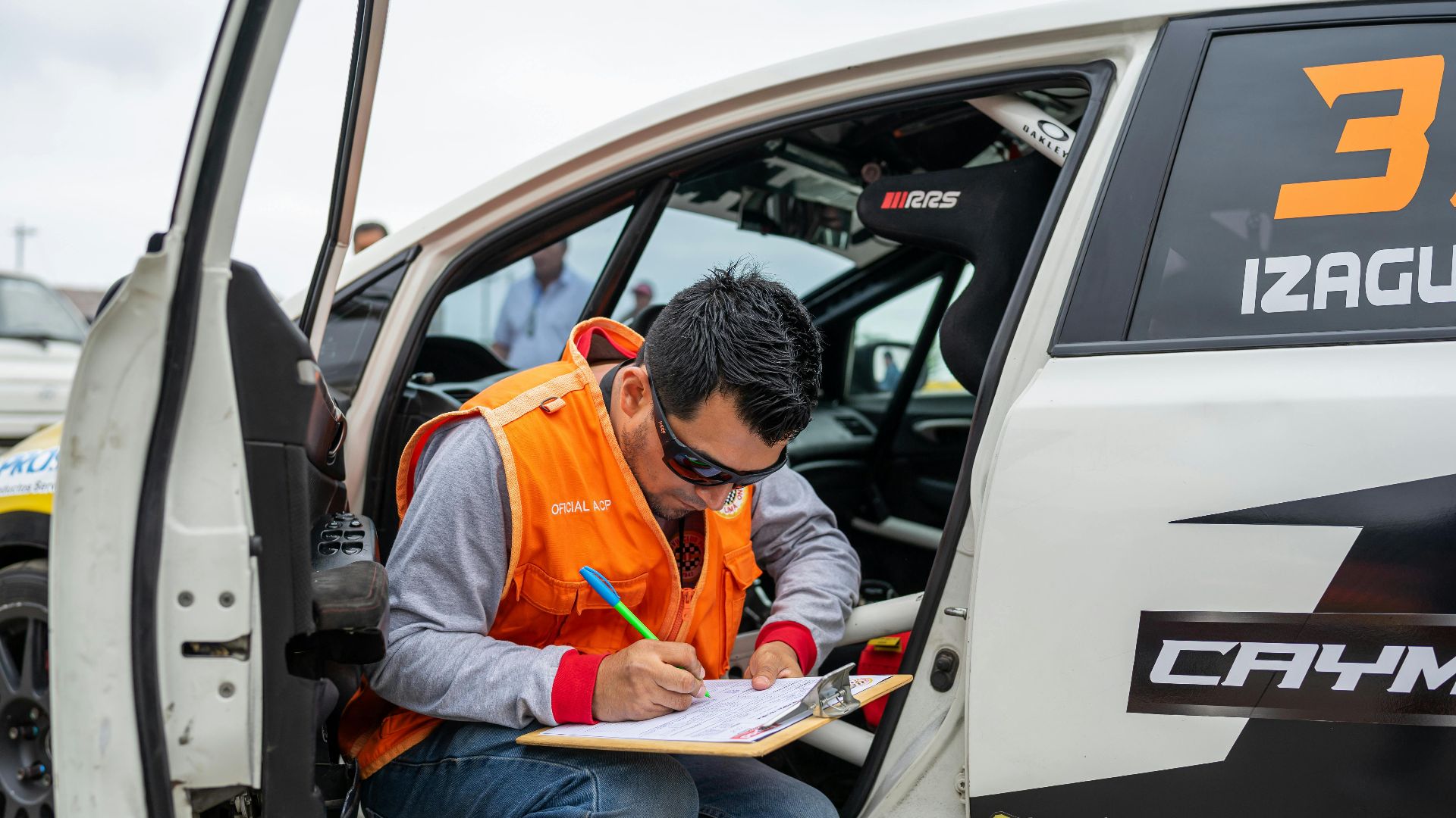 A motorsport official reviews documents while sitting by a rally car during a race event.