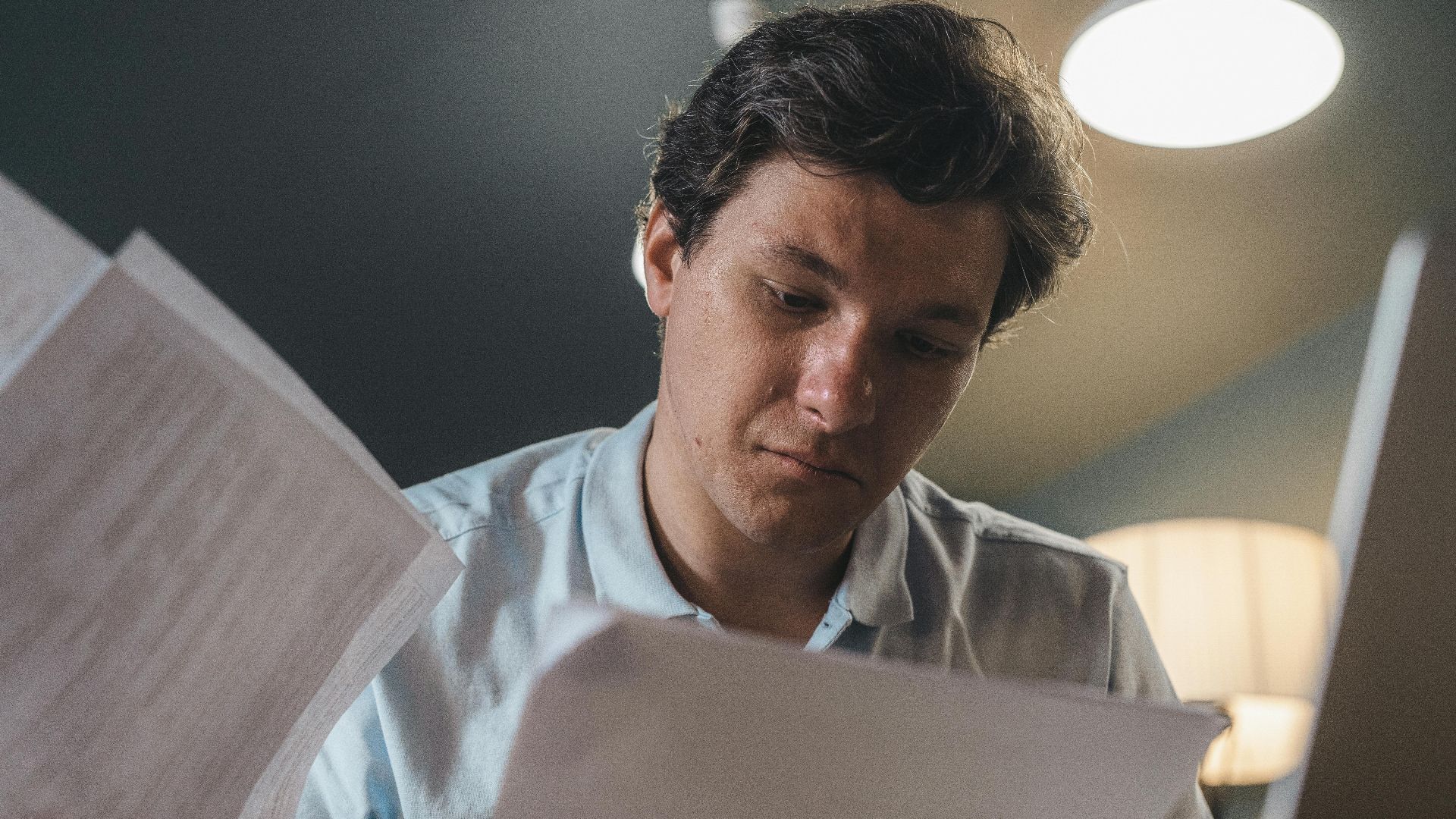 Serious man wearing a light blue shirt intently reading printed papers indoors under warm lighting.
