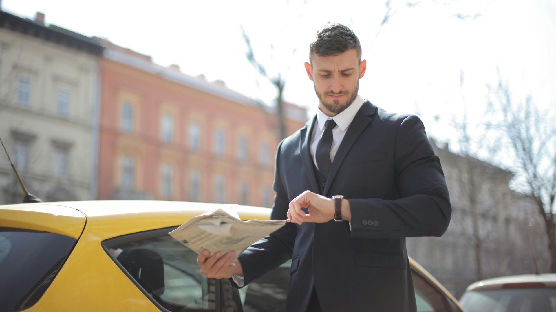 Corporate professional holding newspaper checks watch near a yellow car outdoors.