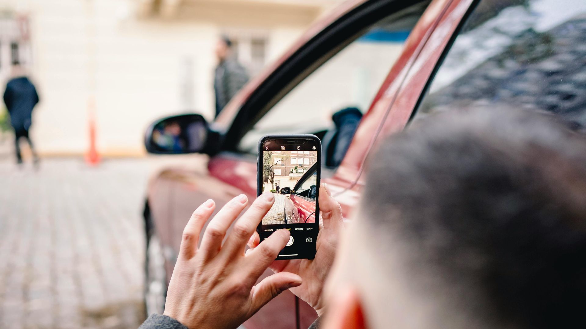 Man using smartphone to take pictures of an urban scene beside a red car.
