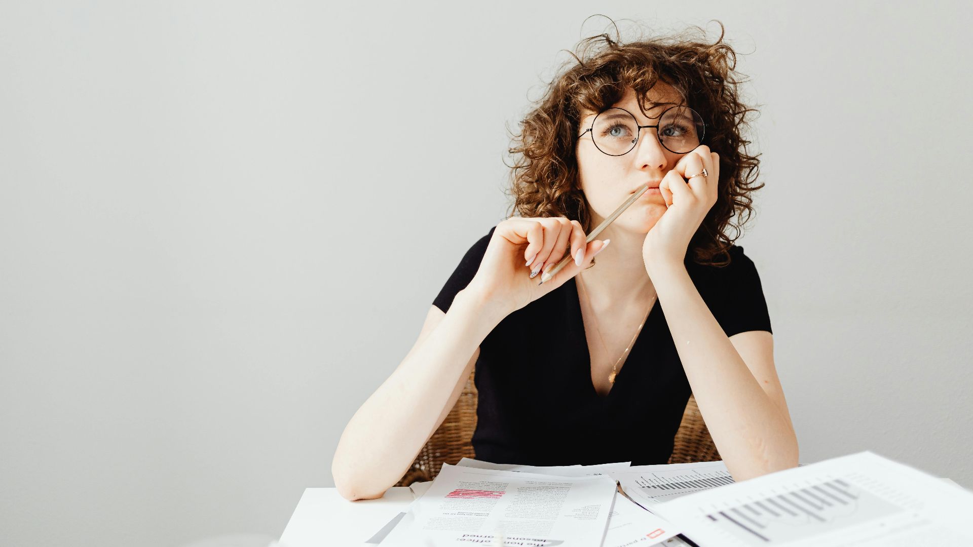 Curly-haired woman with glasses pensively thinking at a desk with documents.
