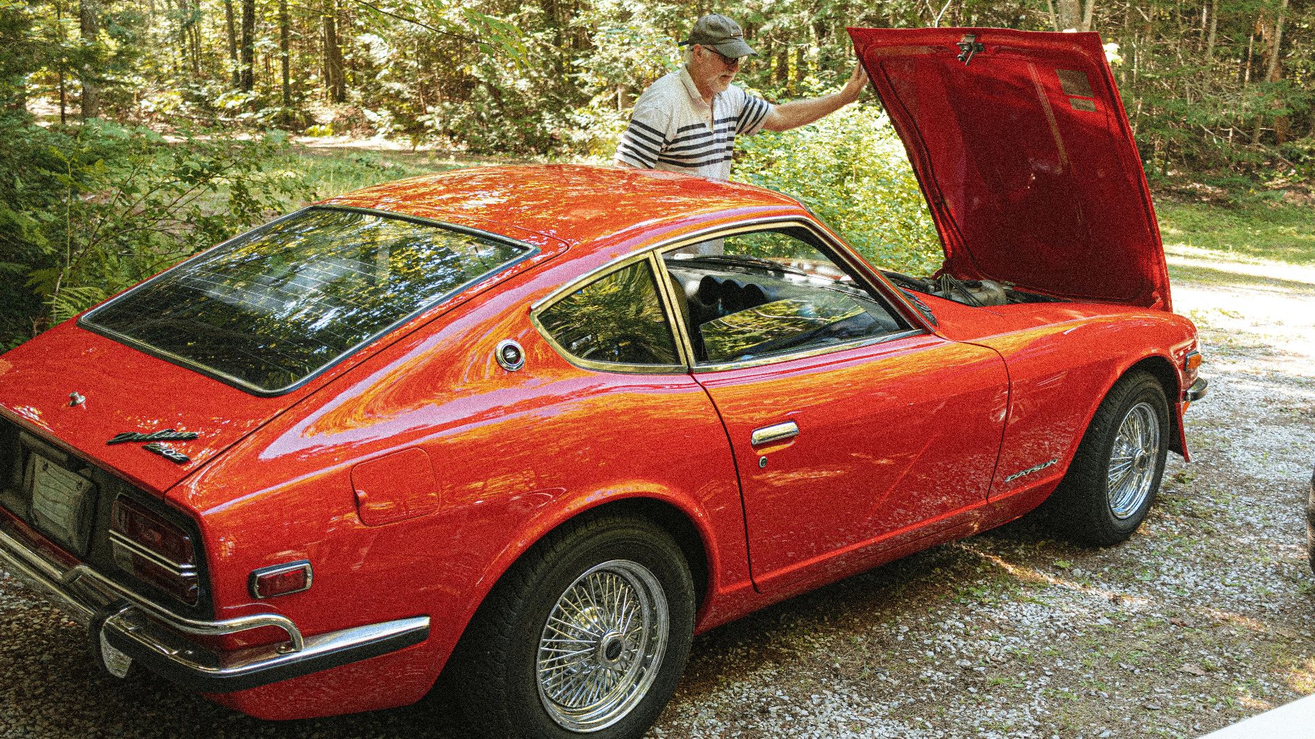 A senior man inspects the engine of a classic red Datsun 240Z parked outdoors.