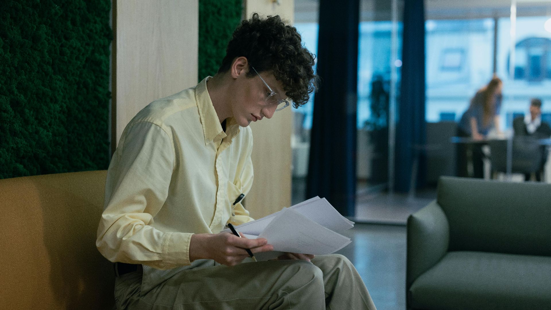 A focused young professional reading documents on a couch in a modern office environment.