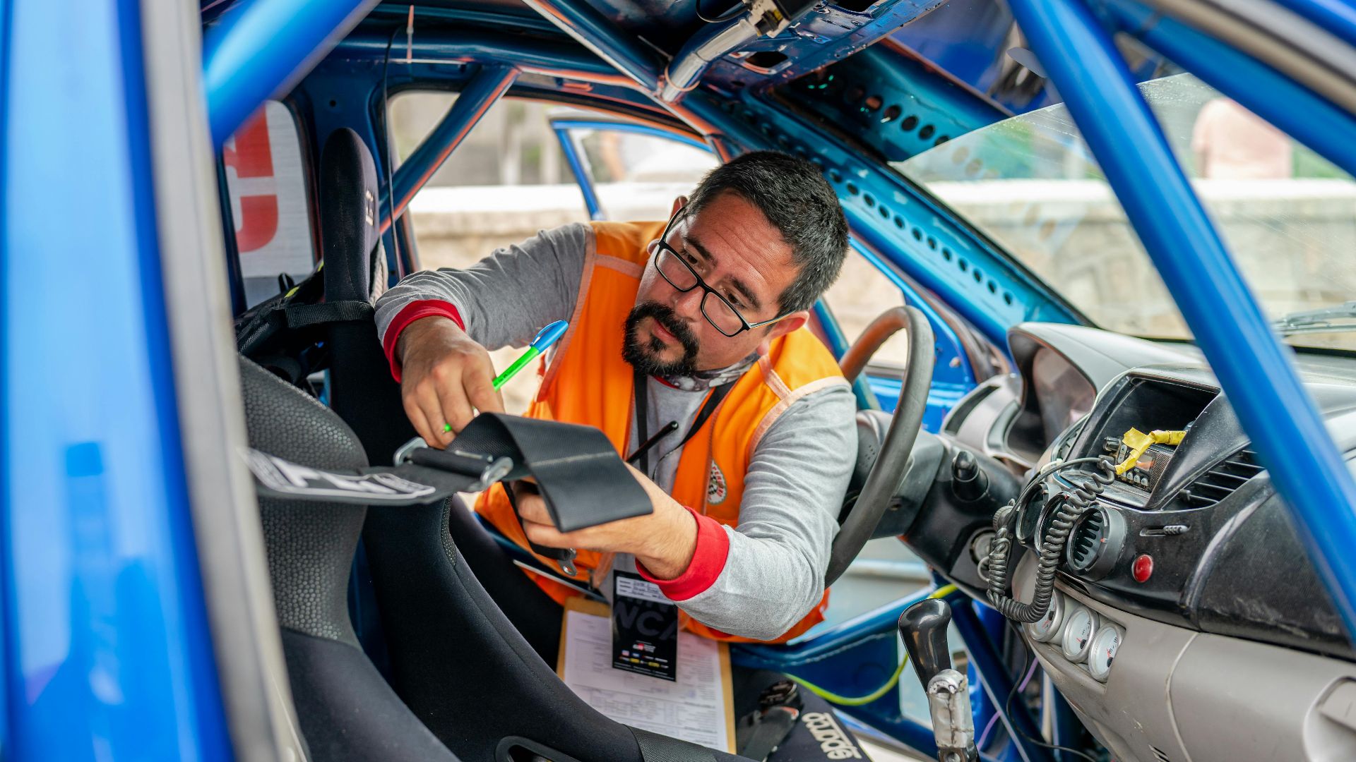 A racing car inspector with eyeglasses checks interior components and safety features in a blue race car.