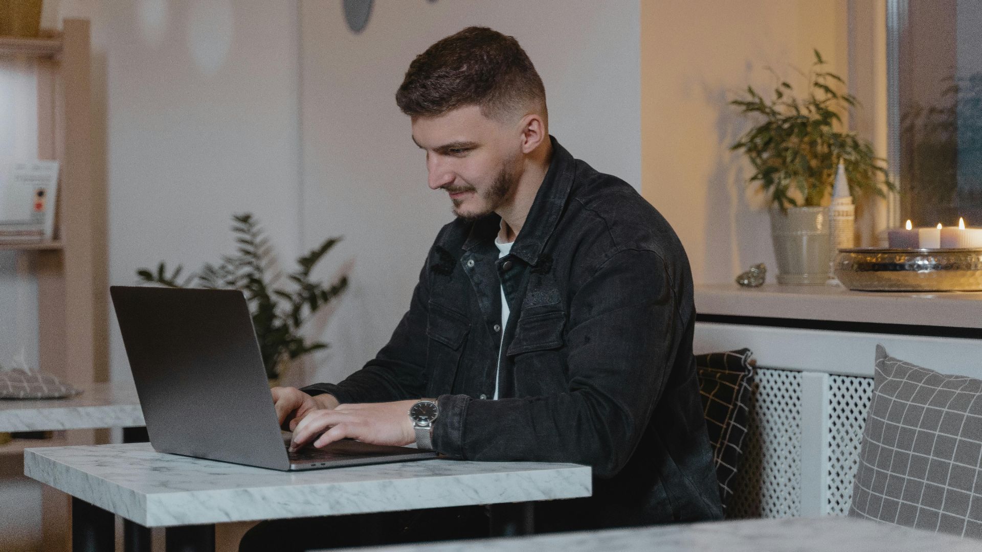 Caucasian man working on a laptop at a cozy indoor setting, surrounded by plants and candles.