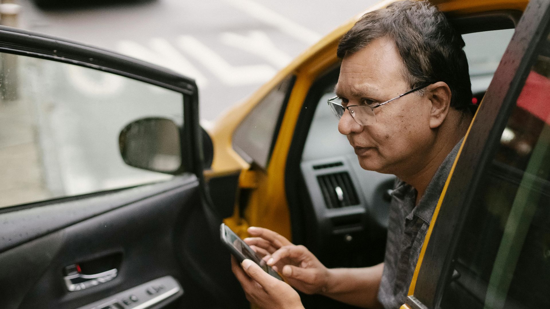 Crop Asian man sitting in parked yellow taxi car with open door and using smartphone while looking away
