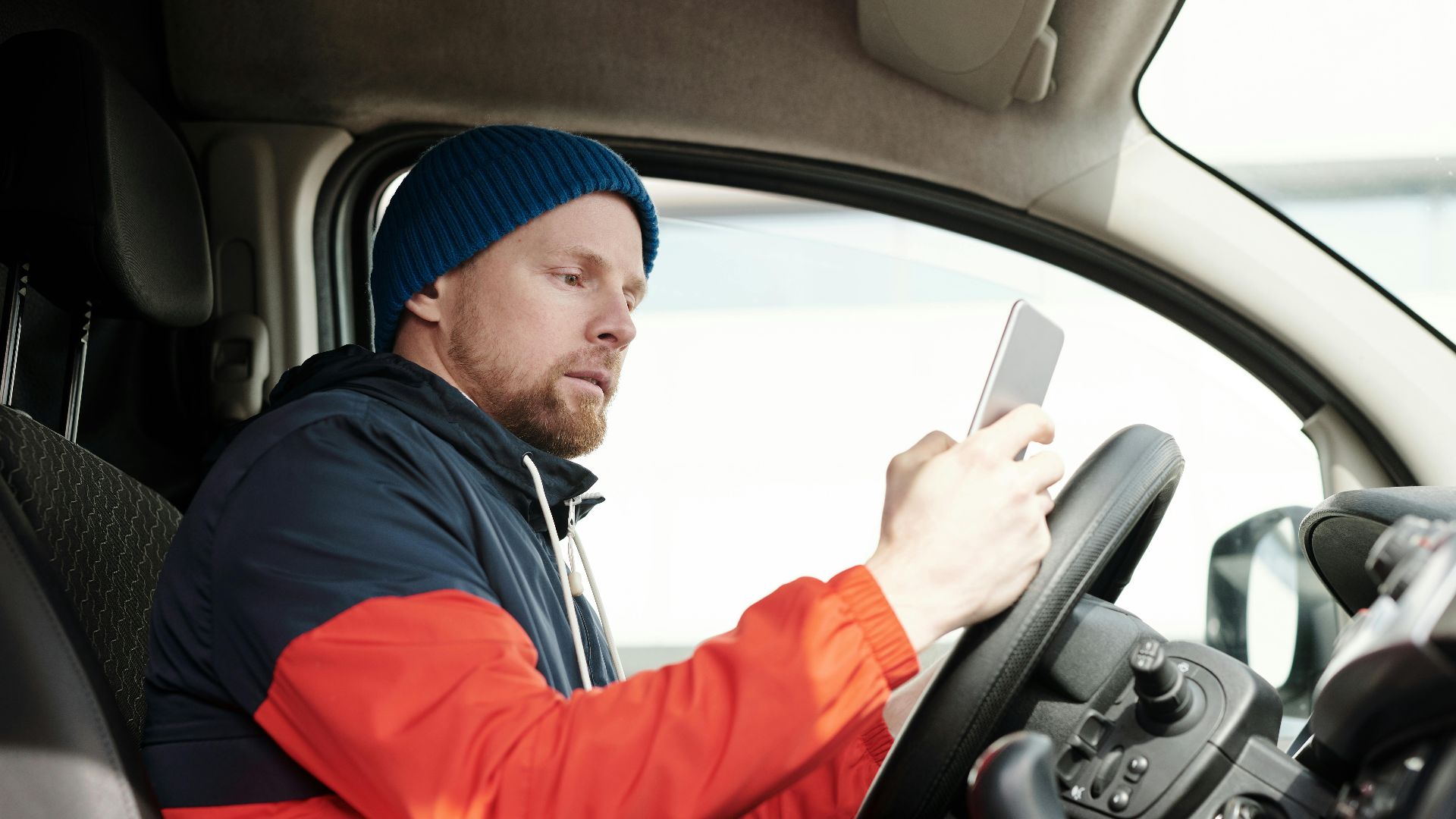 Man in a parked car using a smartphone, wearing a blue beanie and red jacket. Daytime driving scene.