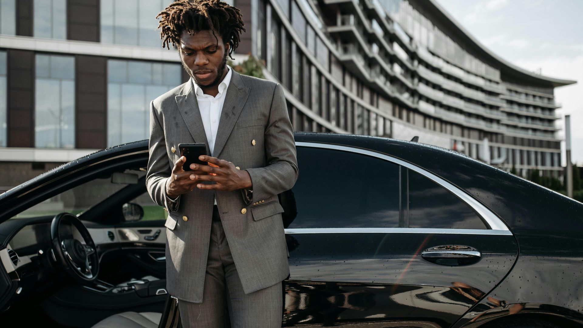 Professional man in a gray suit using his phone next to a luxury car.