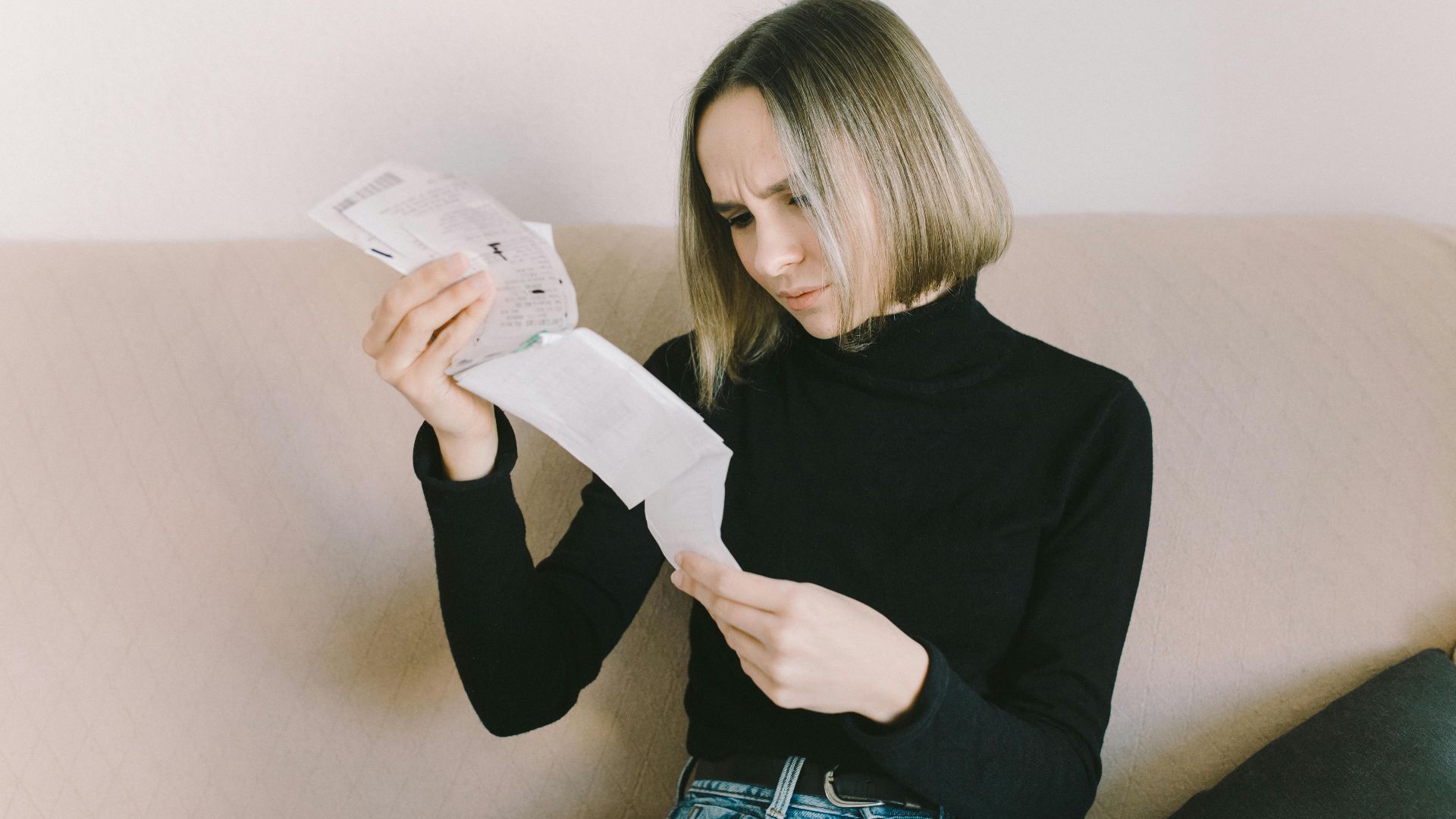 Concerned woman in black sweater examining bills on beige sofa indoors.