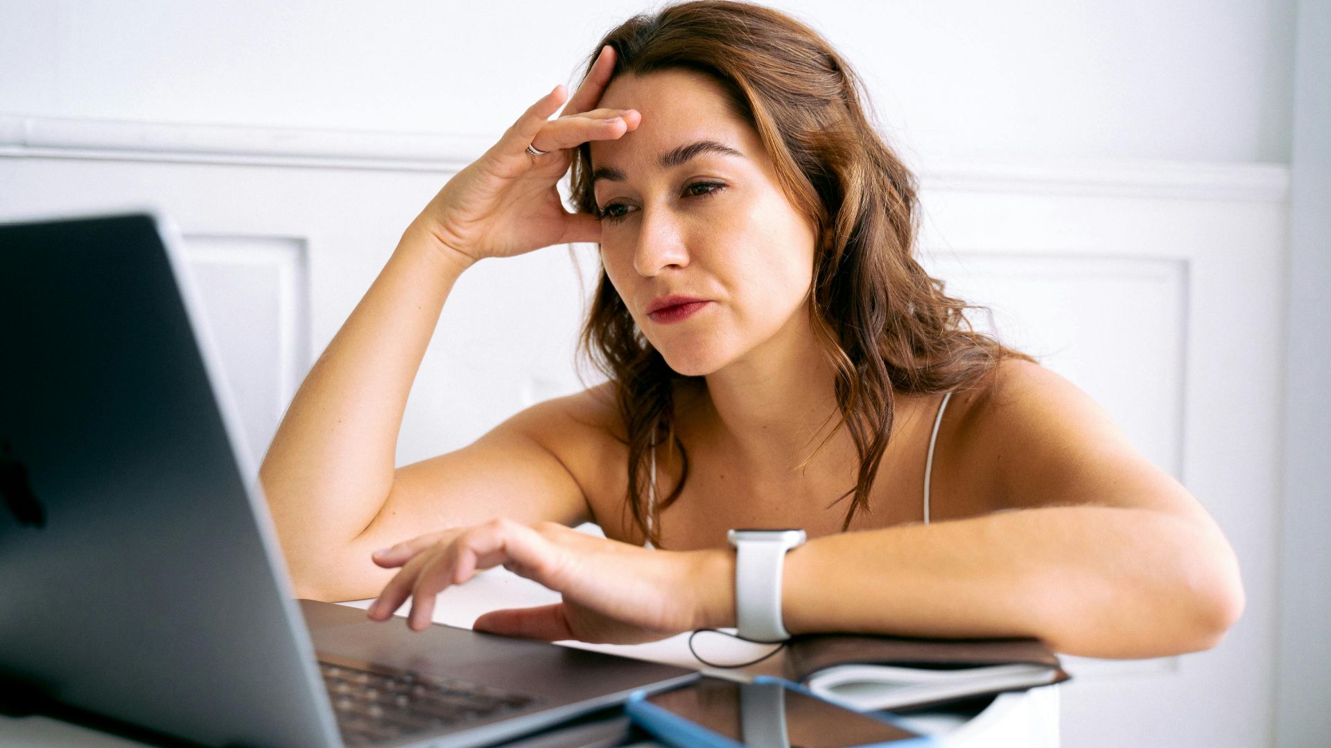 A woman with a frustrated expression, hand on head, working on a laptop.