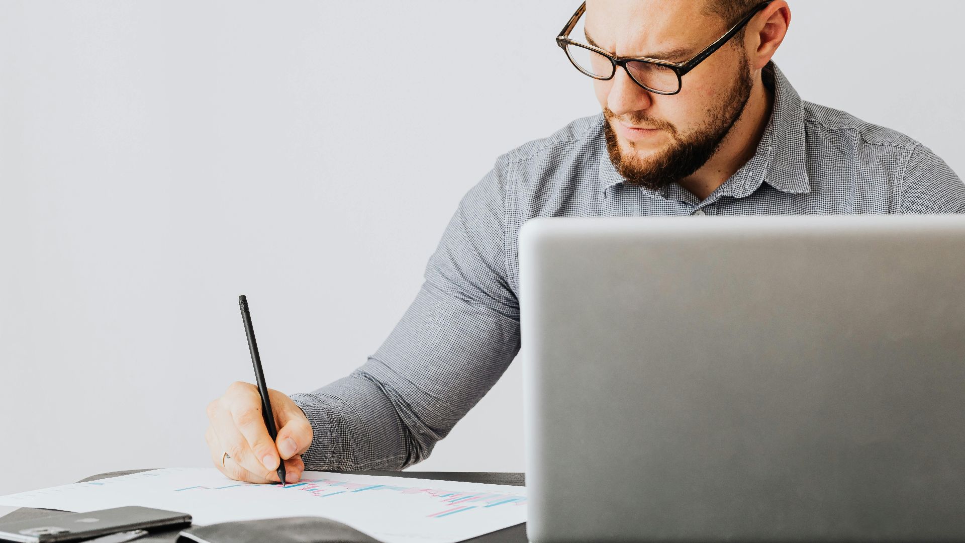 A focused man analyzing financial papers with a laptop in an office setting.