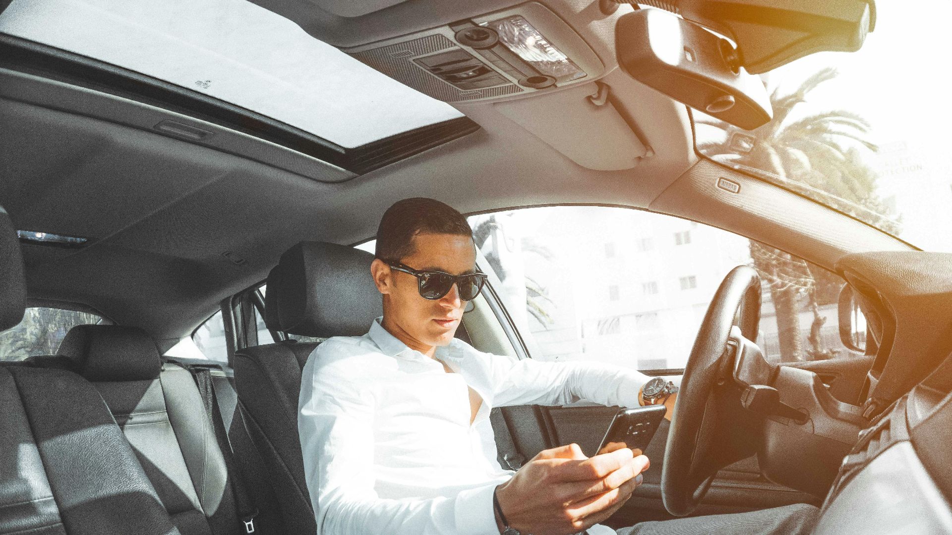 A man wearing sunglasses texts on his phone while driving a car in Morocco.