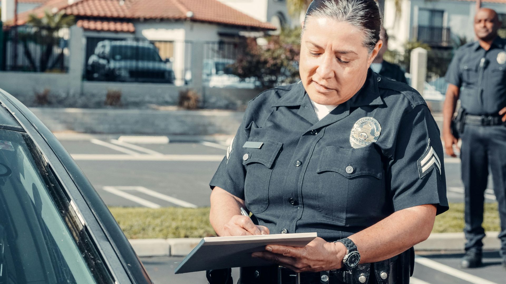 Police officer writing a ticket to a car in a sunny parking lot with a colleague in the background.