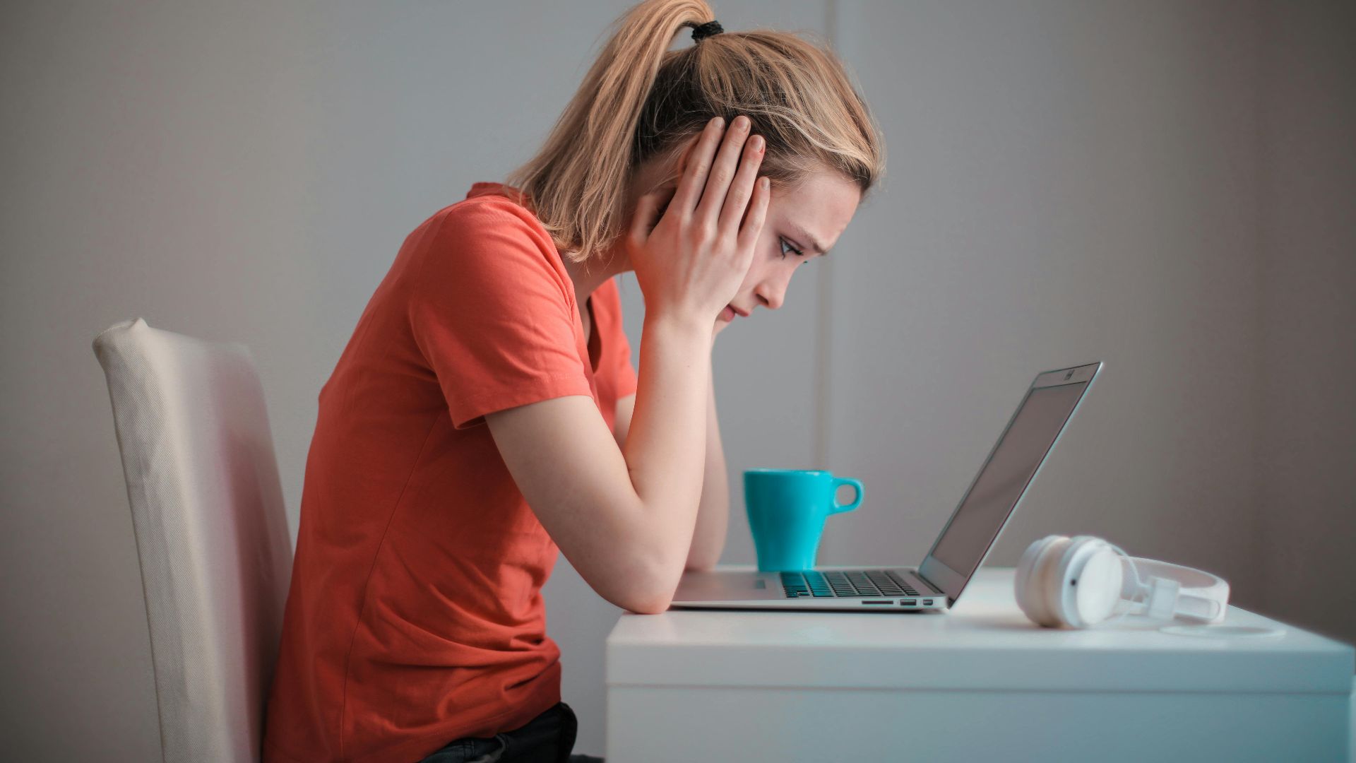 Side view of thoughtful worried female student in casual clothes focusing on screen and trying to solve problem while sitting at table with blue mug of hot drink and headphones and working on laptop at home