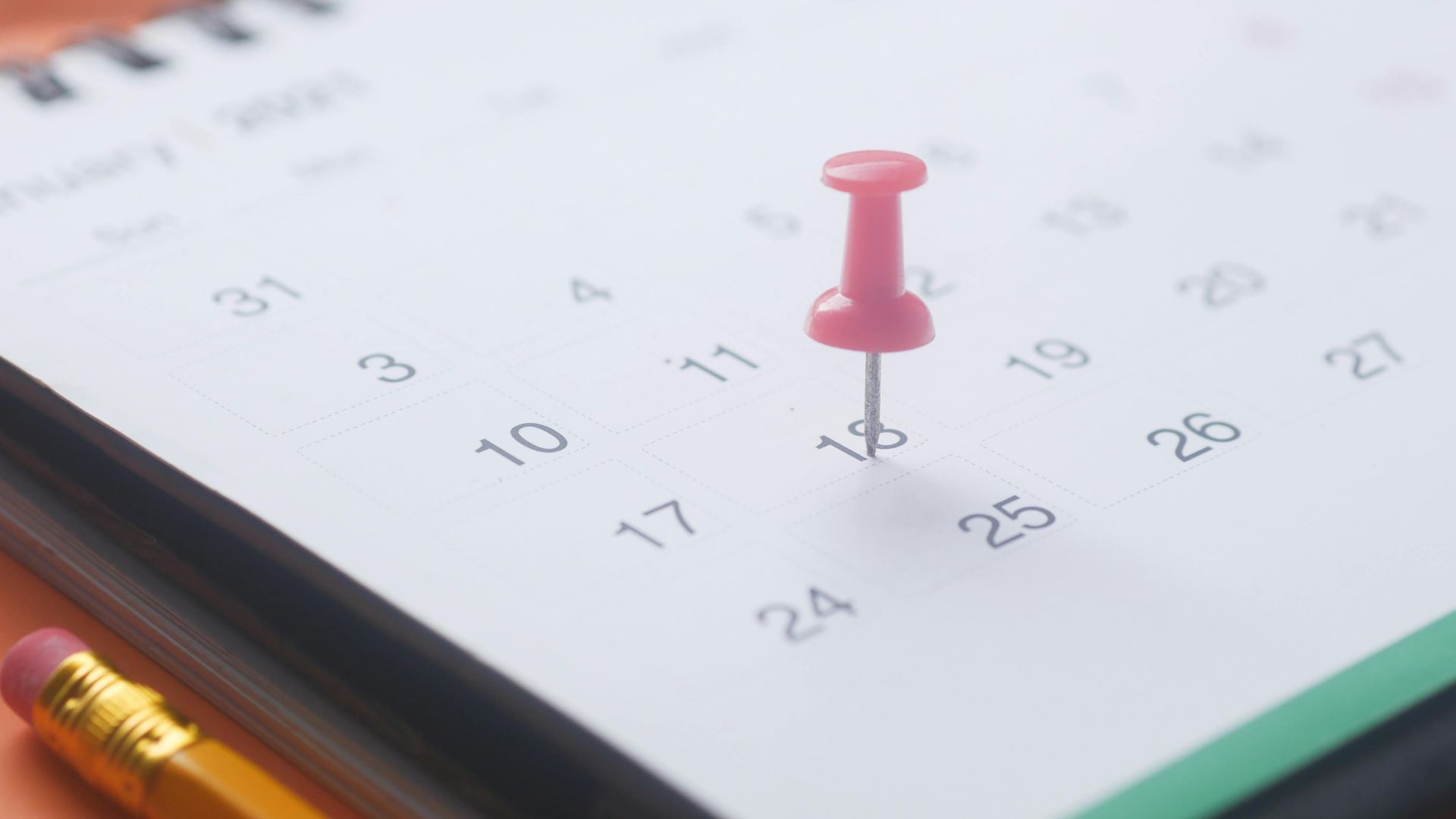 A close-up of a calendar page with a pink push pin, pencil, and binder clip on an orange background.