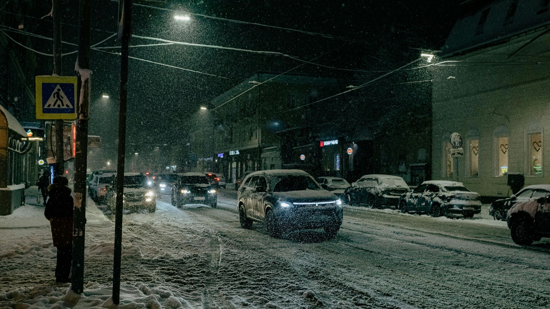 Urban street scene at night with snow, cars, and streetlights illuminating the roadway.