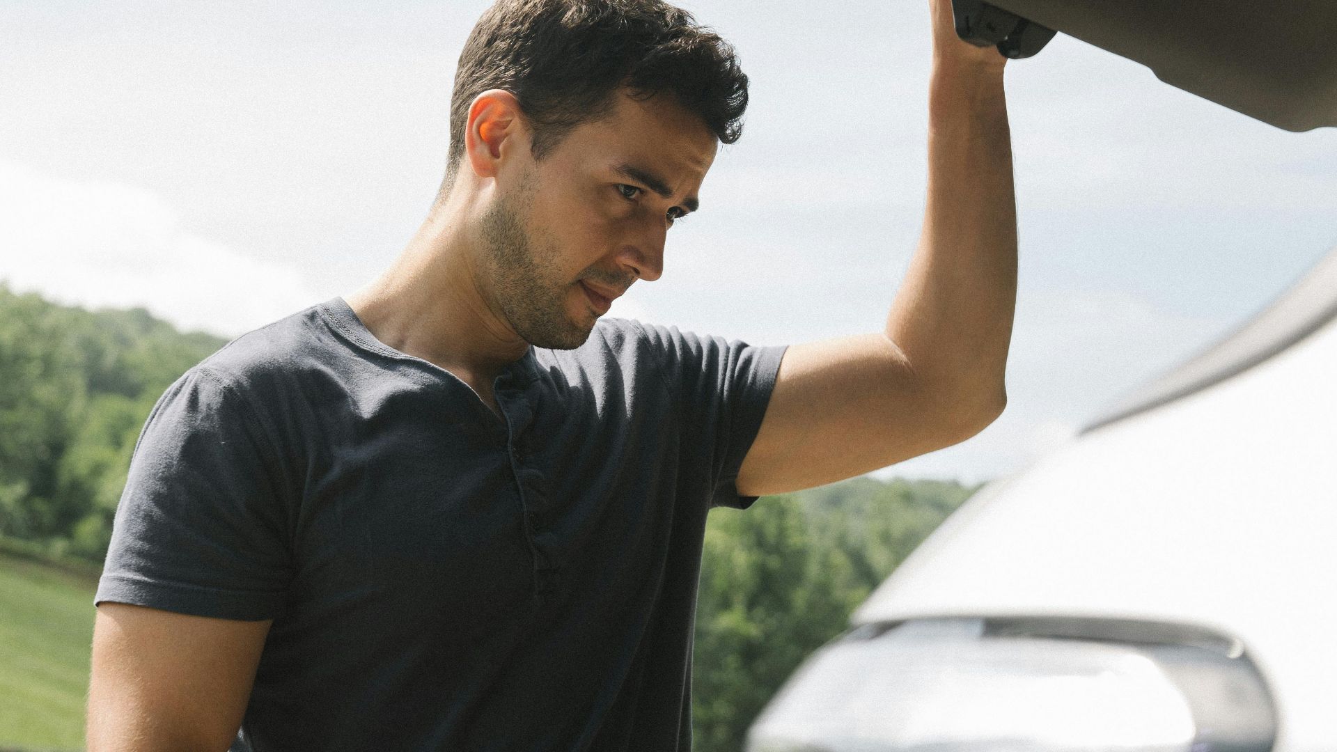 A man examining the open trunk of a car under clear skies with greenery in the background.