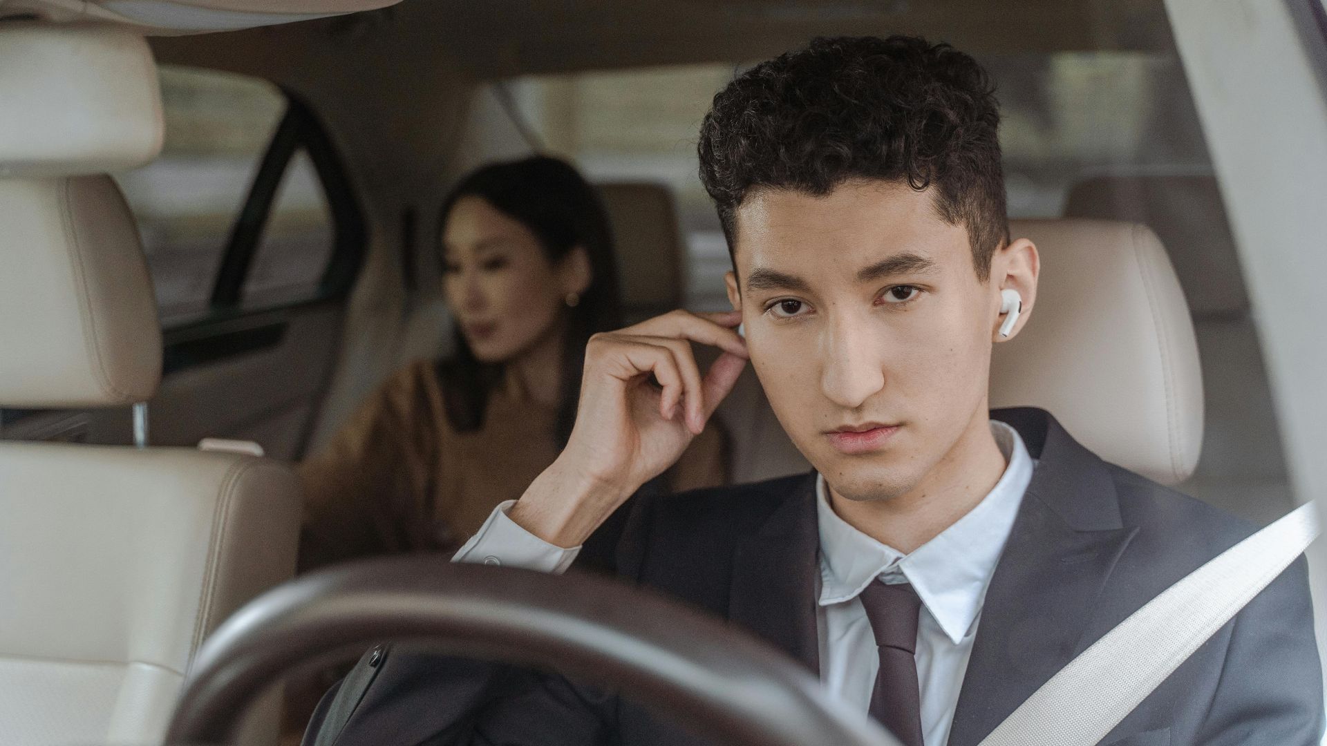 A professional driver in a suit with AirPods driving a female passenger in a car.