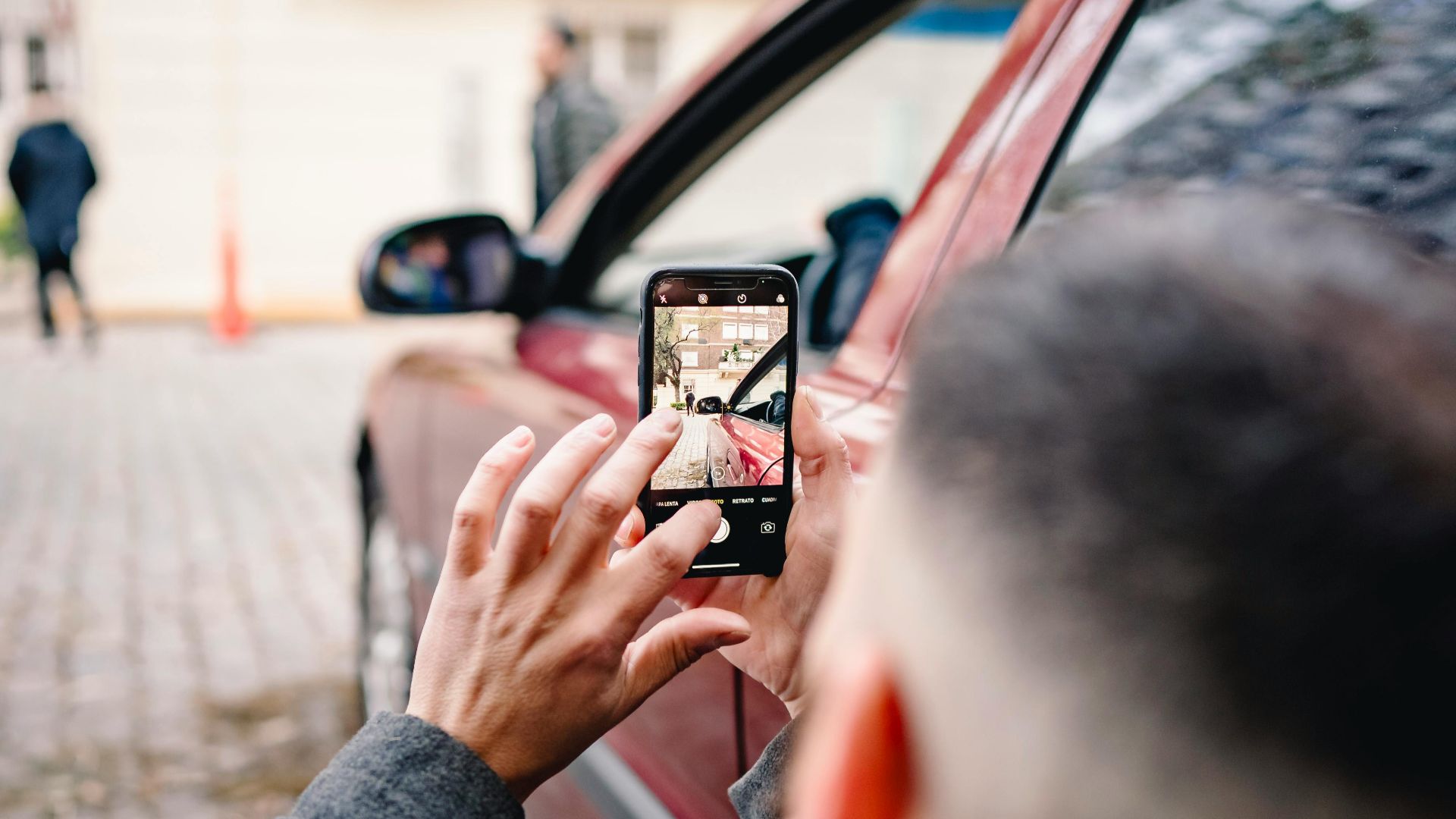 Man using smartphone to take pictures of an urban scene beside a red car.