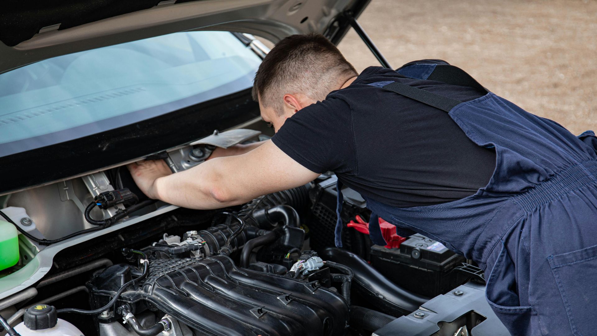 A mechanic in overalls works on a car engine outdoors, showcasing automotive maintenance.