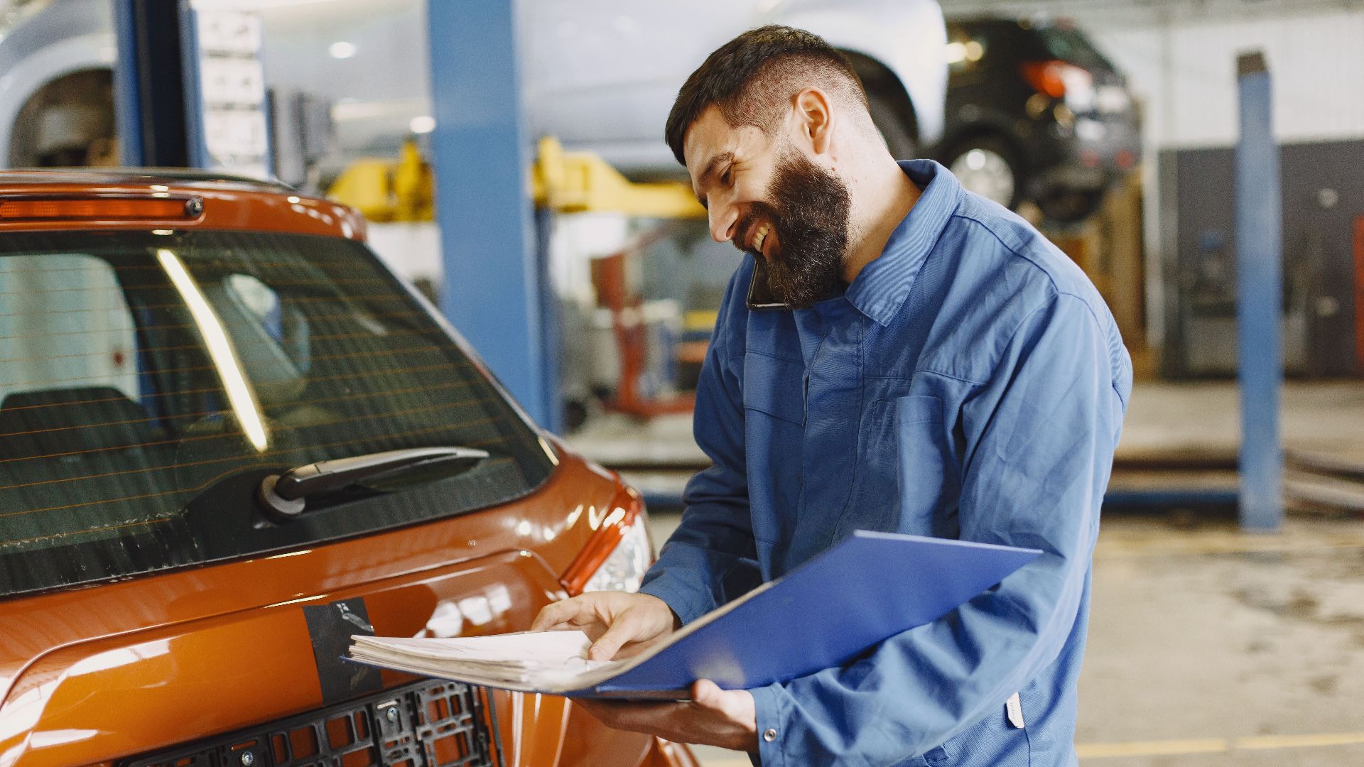 Smiling auto mechanic inspects a car in a service garage, wearing coveralls and holding documents.