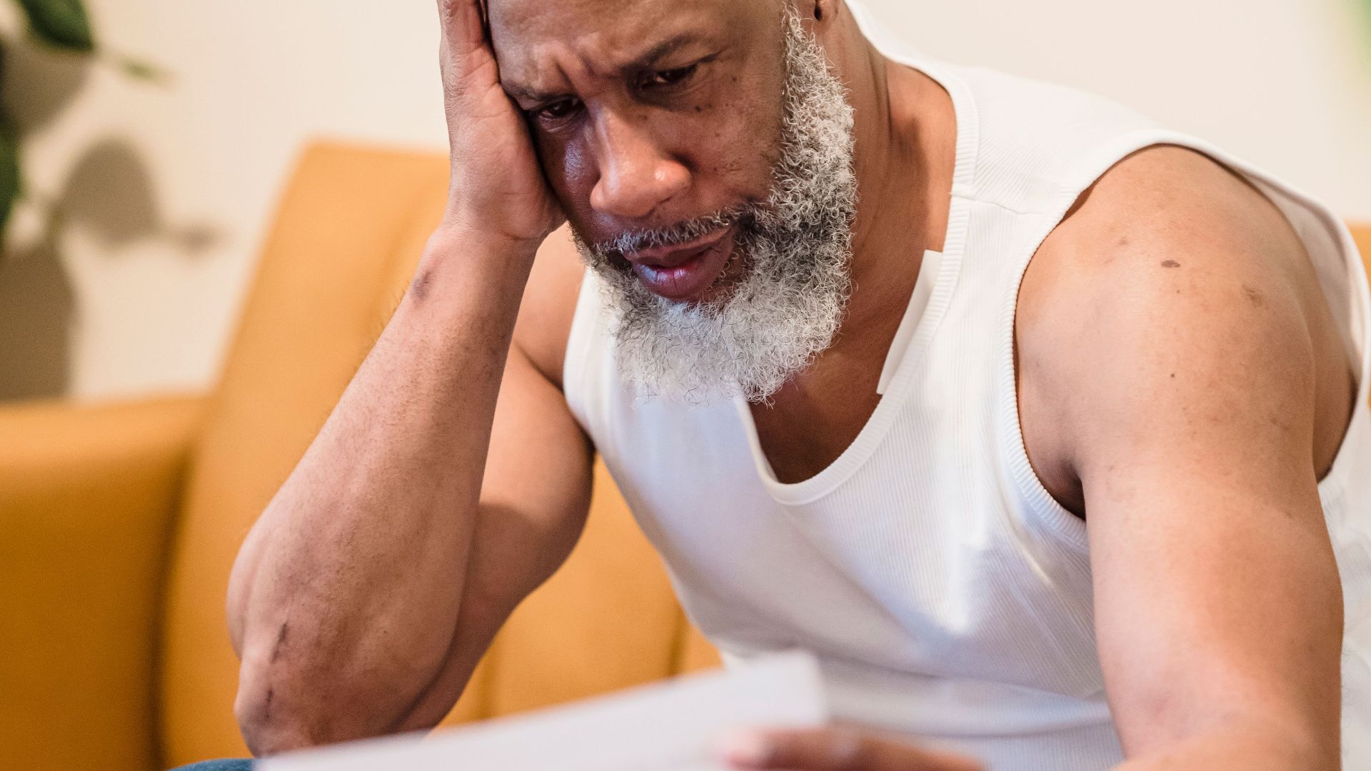 A worried man sitting on a sofa examining documents, expressing anxiety.