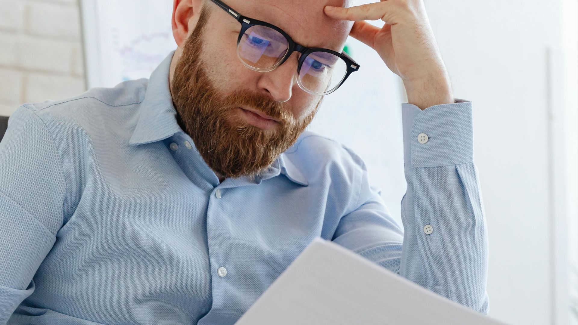 Caucasian businessman with beard and glasses reviewing documents in a modern office setting.