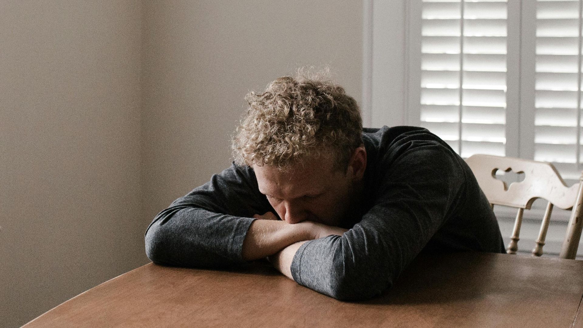 An adult man with emotions of sadness and anxiety sitting alone at a wooden table near windows.