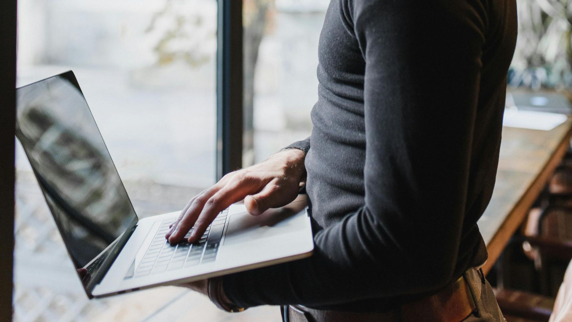 Adult man using a laptop indoors by a large window, focused on work.