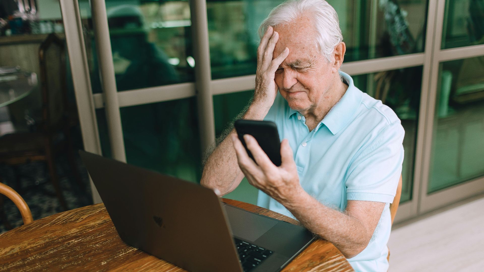 Elderly man frustrated by smartphone and laptop technology indoors at home.
