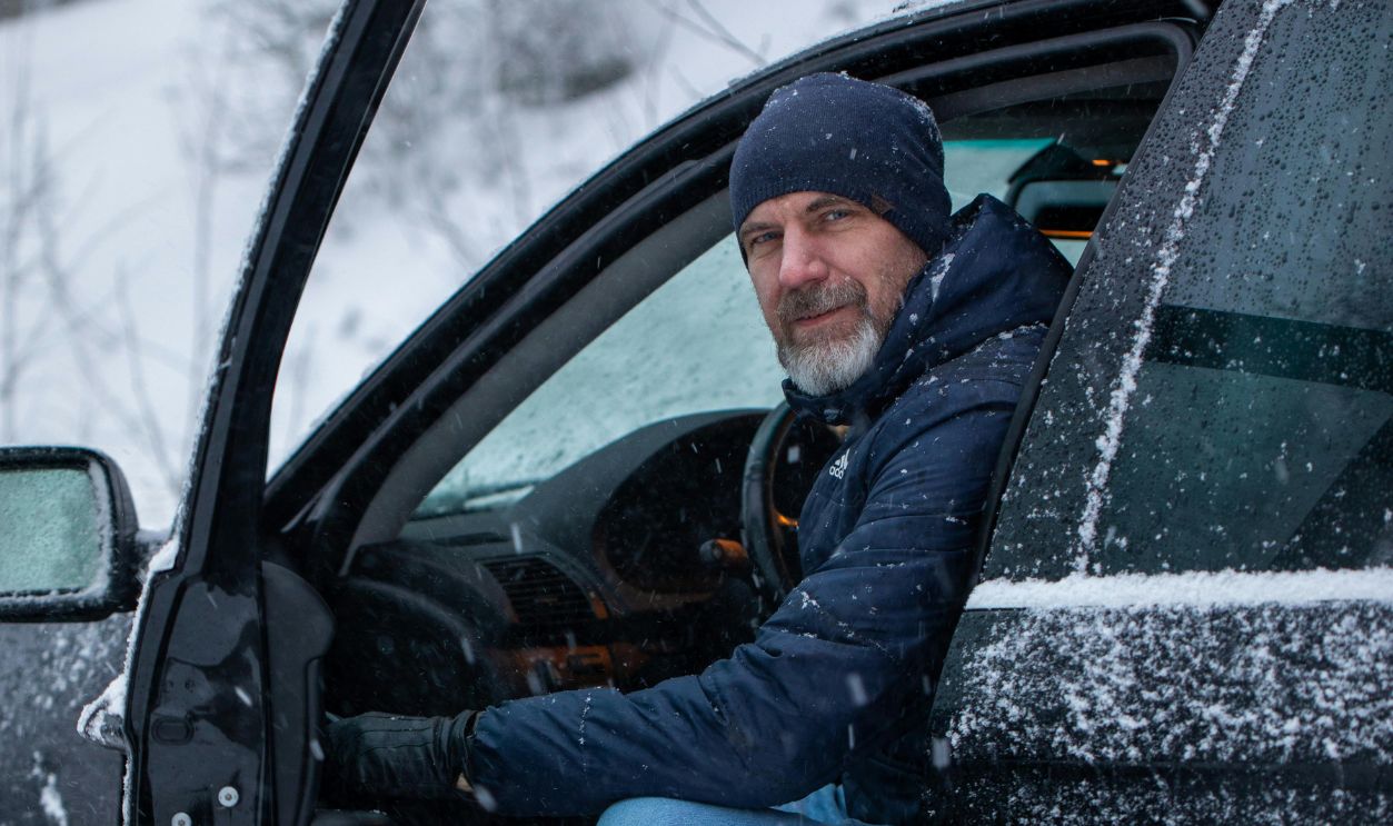 A man in winter attire sits in a car doorway amid falling snow