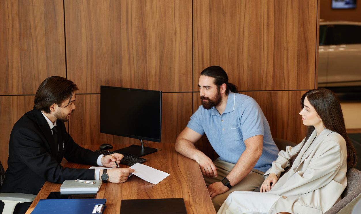 Shutterstock-2513248373, Side view of male car broker at desk making deal with couple buying new vehicle, discussing terms and conditions of contract at office of dealership center