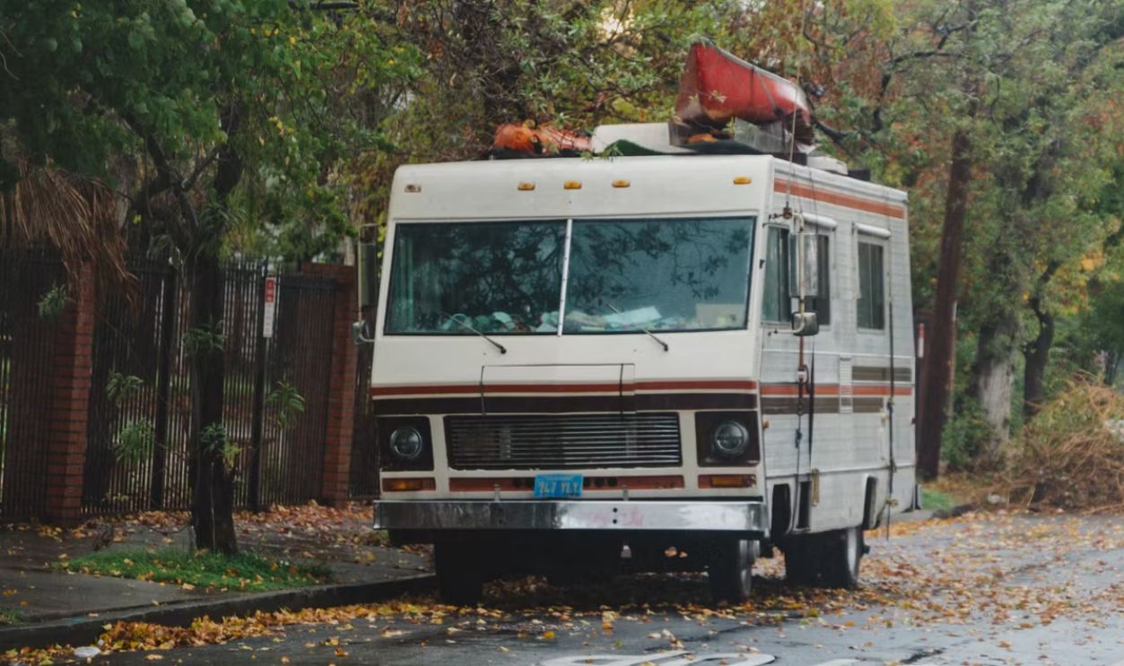 A vintage RV with a red canoe on top parked on a tree-lined street in an autumn setting