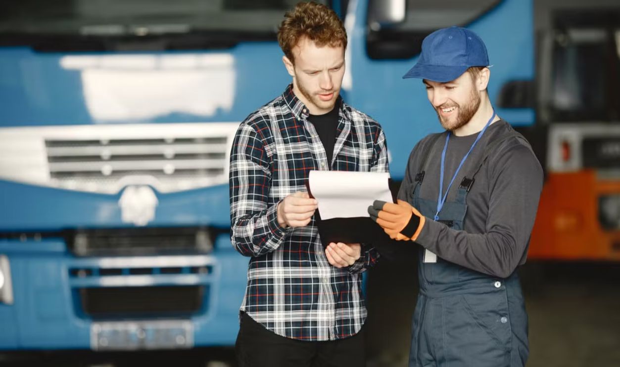 Two men in discussion over vehicle maintenance documents in a garage setting with trucks in the background