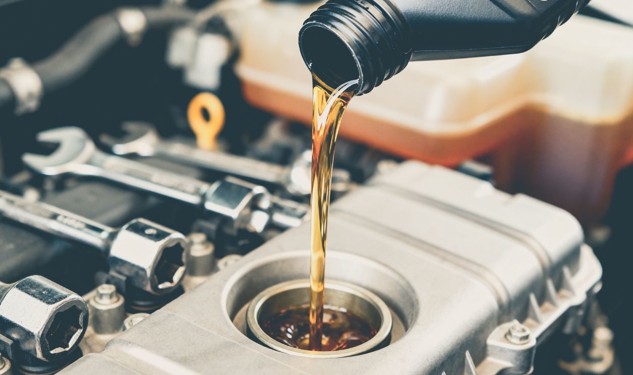 Shutterstock-2726414421, A close-up view of golden engine oil being poured from a black bottle into the oil reservoir of a car engine, with various engine components visible in the background.