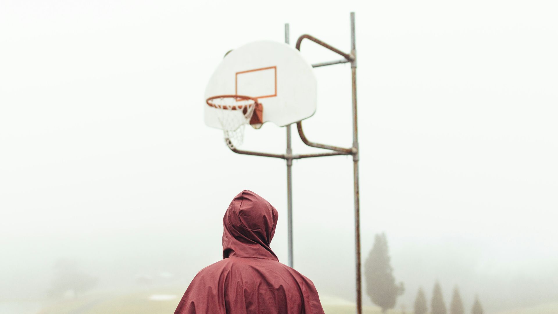 person wearing maroon raincoat standing under white and grey basketball hoop during foggy daytime