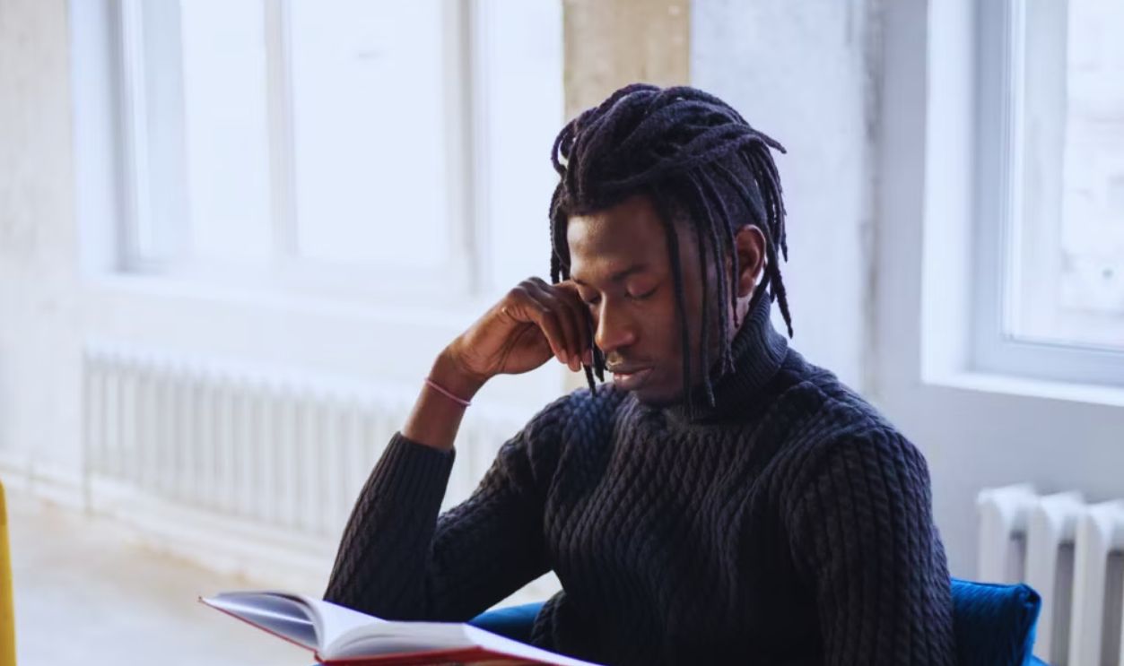 A young man with dreadlocks attentively reading indoors by a bright window