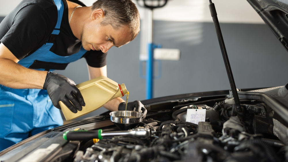 The mechanic is pouring oil into the engine. Pouring fresh oil to car engine, oil change to auto. Caucasian man in blue overalls pouring oil from plastic container.