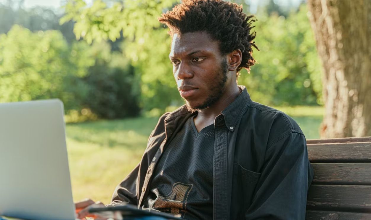 Young man with afro hair working on a laptop while sitting in a sunny park.