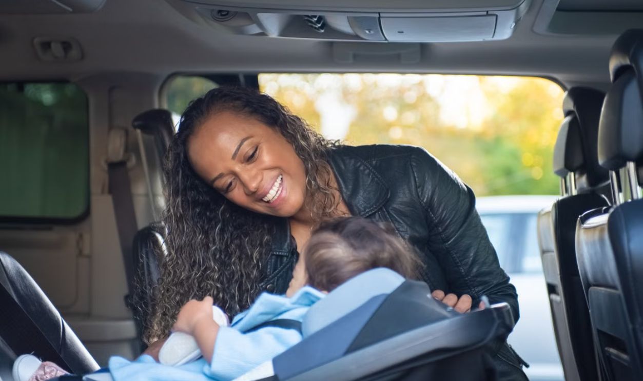 A joyful mother smiles at her baby in a car seat inside a vehicle.