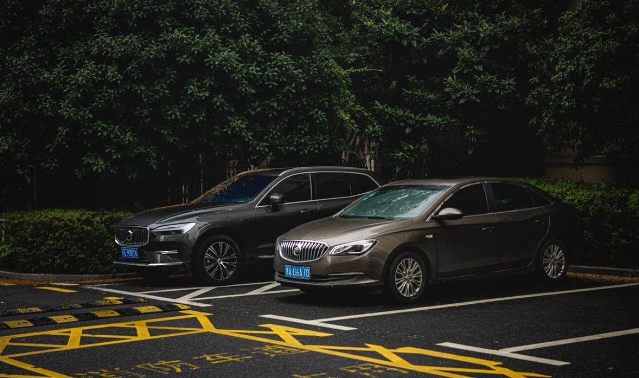 Two cars, an SUV and a sedan, parked side by side in a shady urban lot.