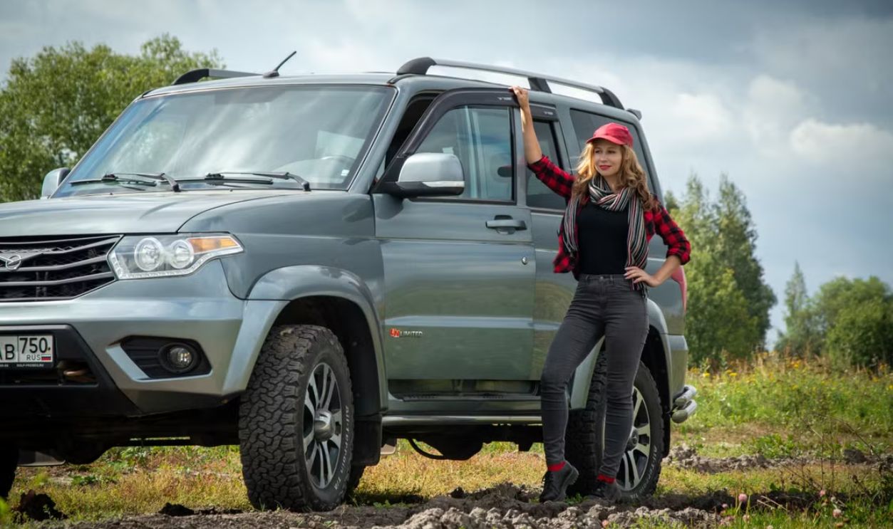 A stylish woman stands confidently beside an off-road SUV in a natural landscape.
