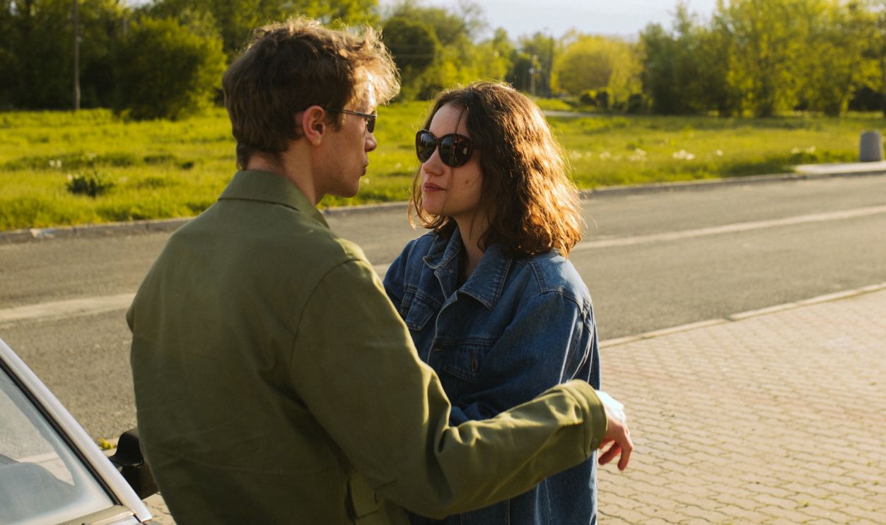 Young couple outdoors, leaning on a car, embracing and enjoying a sunny day.