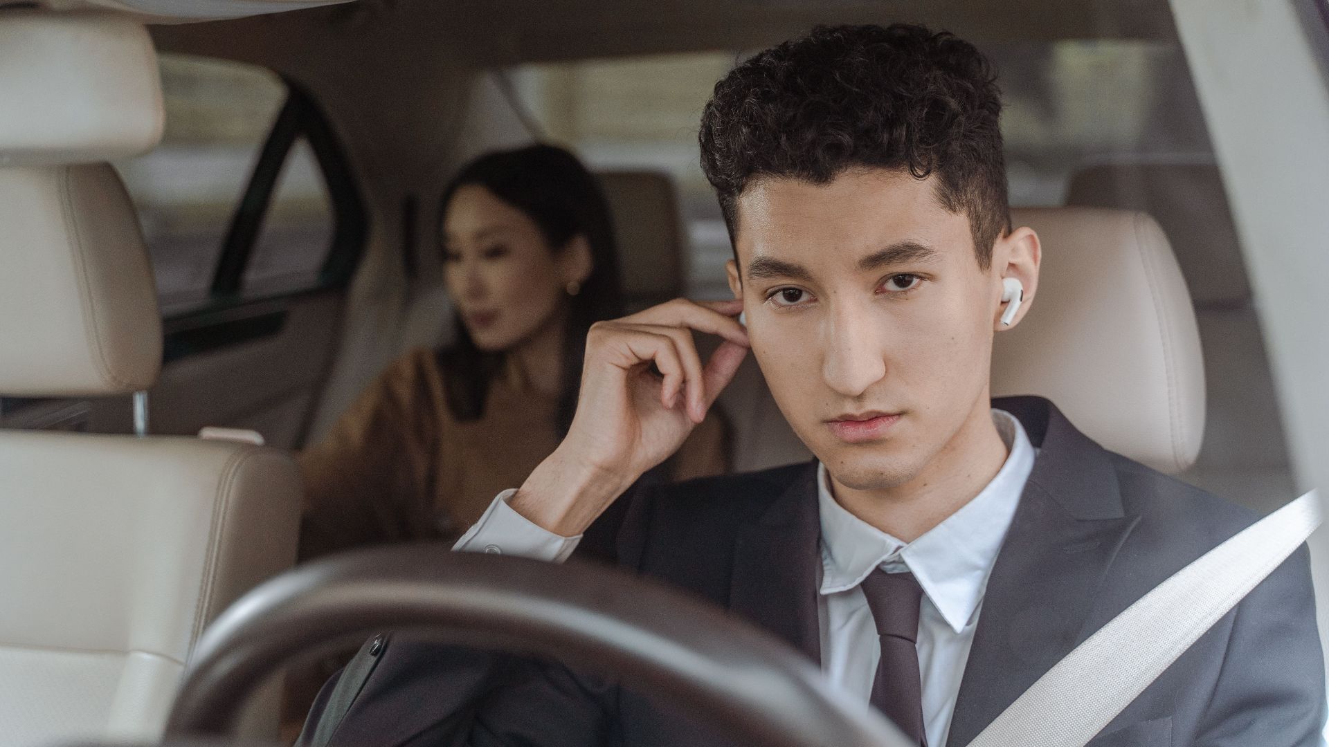 A professional driver in a suit with AirPods driving a female passenger in a car.