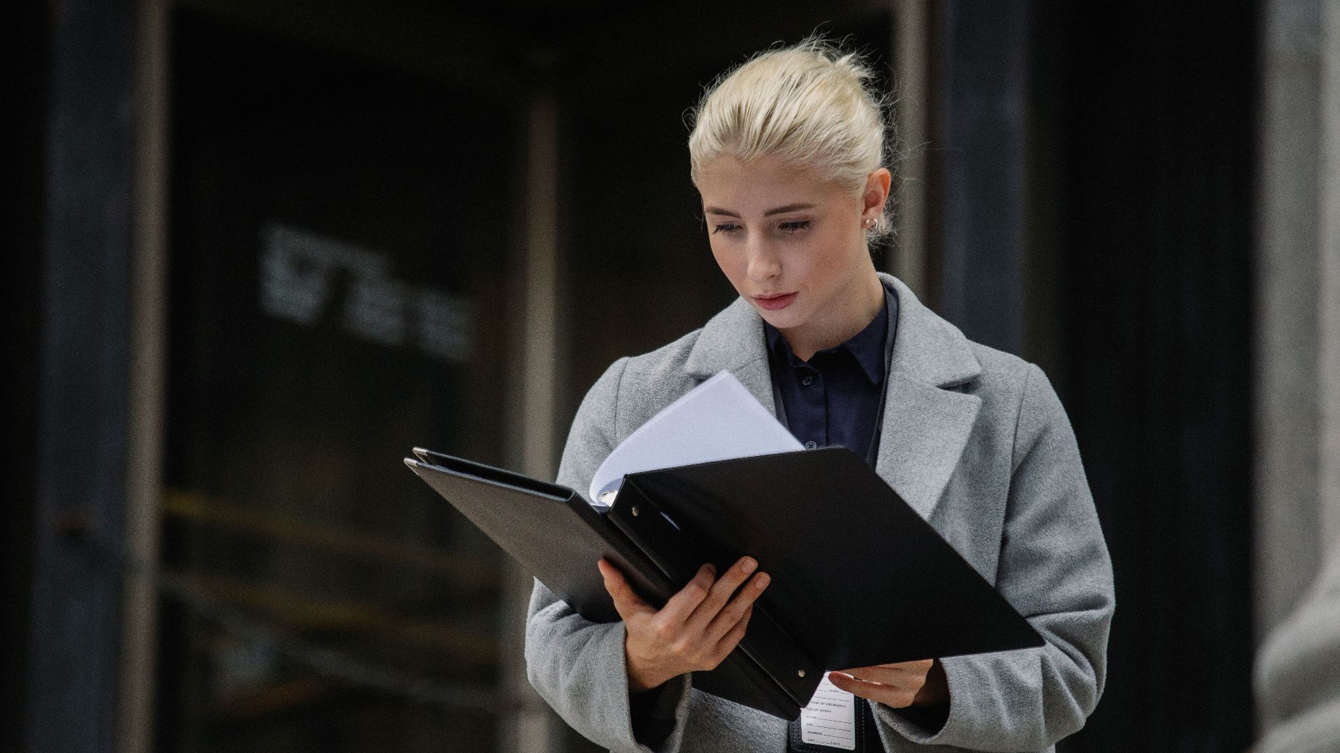 Focused young businesswoman in formal clothes standing outside modern building and reading notes in folder