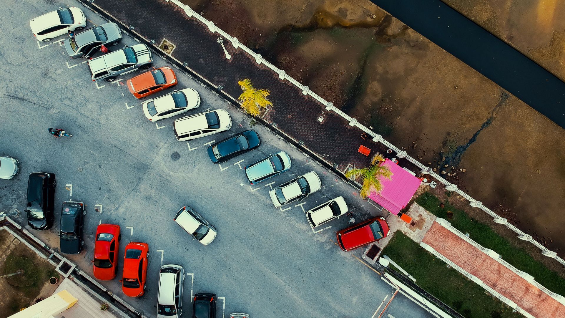 Aerial shot of a parking lot with cars and a pink building, next to a waterway.