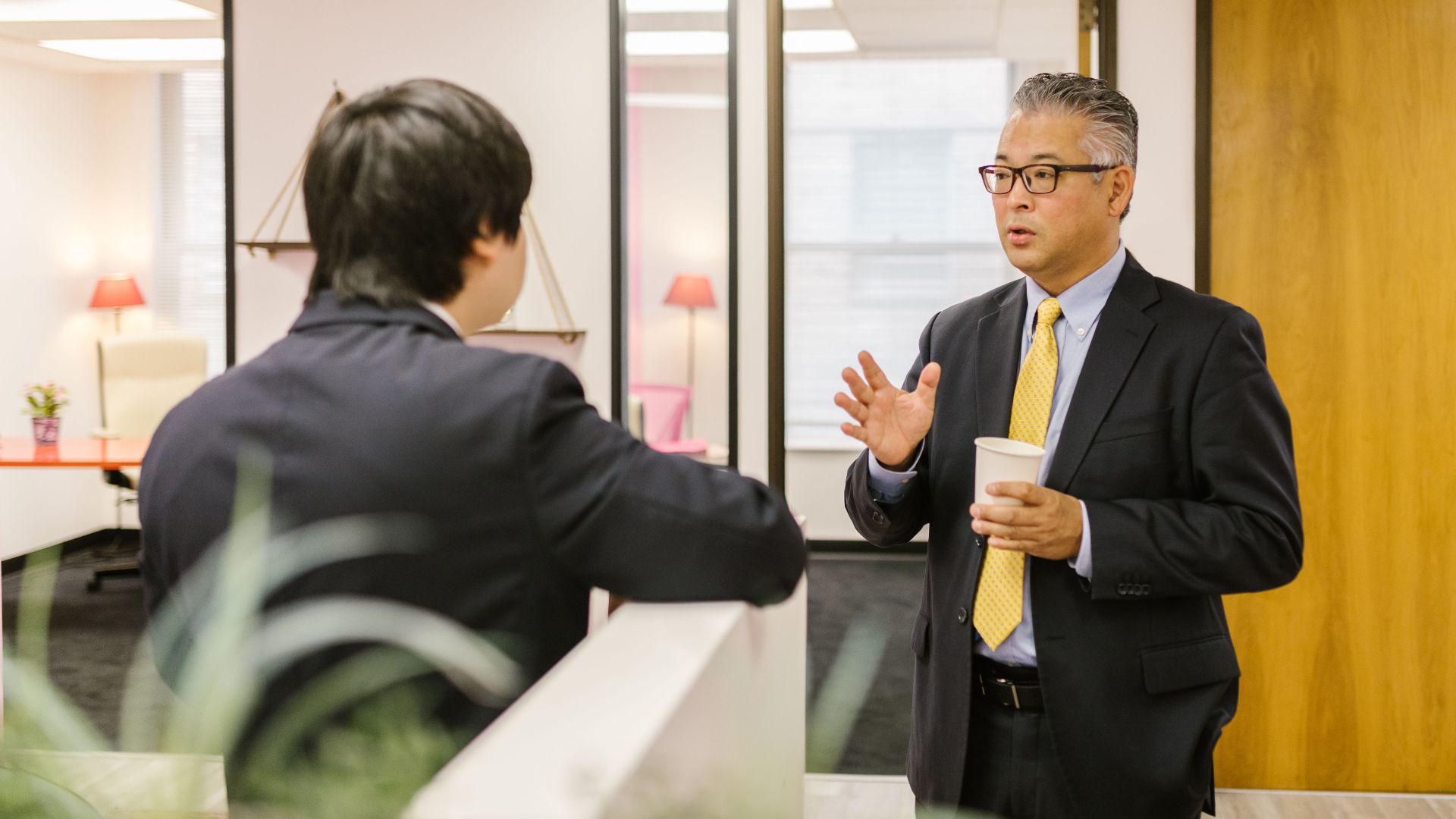 Two businessmen in formal attire having a conversation in an office setting.