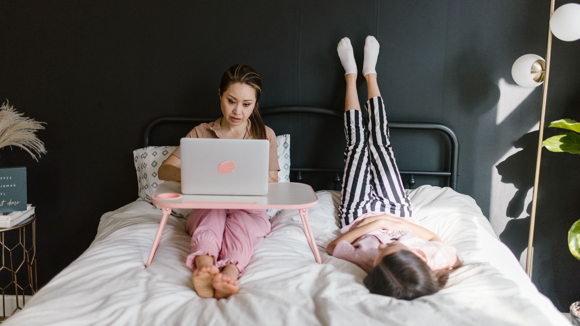 Woman working remotely on laptop in bed, child playing nearby, embracing new work-from-home culture.